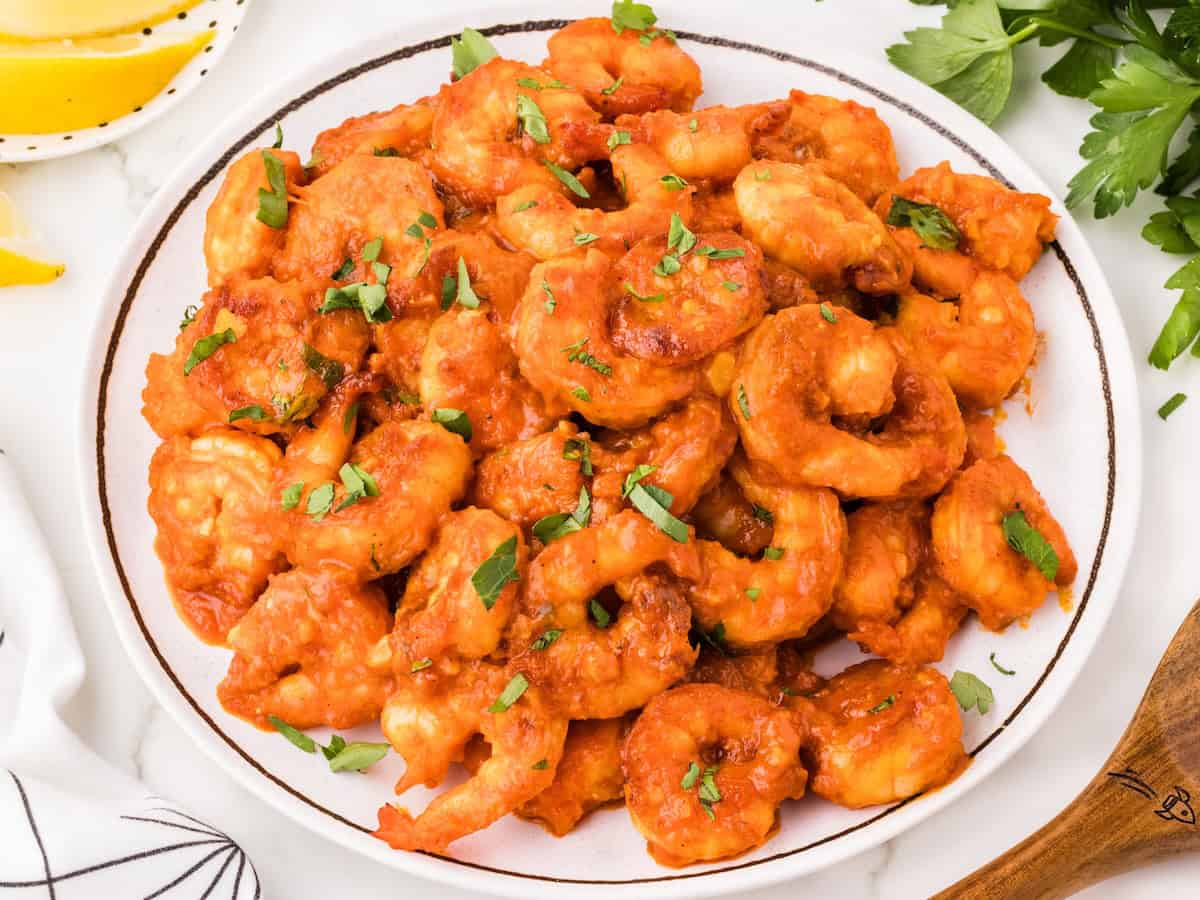 A plate of Spicy Garlic Shrimp in a vibrant red sauce, garnished with chopped fresh herbs, sits on a white table near lemon wedges and green parsley.