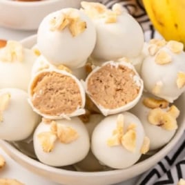 Bowl of banana pudding truffles coated in white chocolate and topped with banana chips, surrounded by vanilla wafers and bananas.