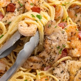 Close-up of creamy chicken and shrimp carbonara with spaghetti, seared chicken pieces, shrimp, and bacon tossed in a rich sauce, garnished with chopped parsley and served with metal tongs.
