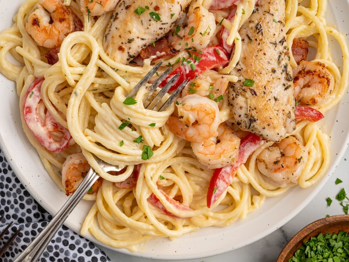 Top view of creamy chicken and shrimp carbonara with spaghetti, seasoned chicken strips, shrimp, bacon, and red pepper slices in a rich sauce, garnished with parsley and served with a fork on a white plate.