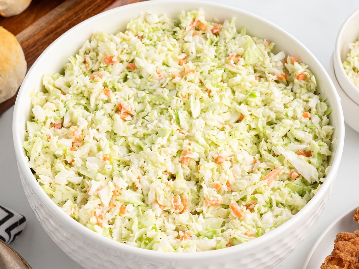 A large white bowl filled with creamy KFC Copycat Coleslaw made of finely shredded cabbage and carrots, set on a white surface with part of some bread rolls and other dishes visible nearby.