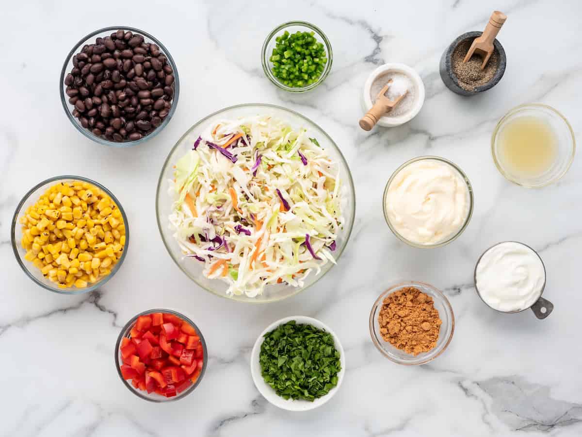 A large central bowl of taco coleslaw surrounded by small bowls of black beans, corn, red bell pepper, chopped jalapeños, cilantro, spices, mayonnaise, sour cream, and lime juice on a marble surface.