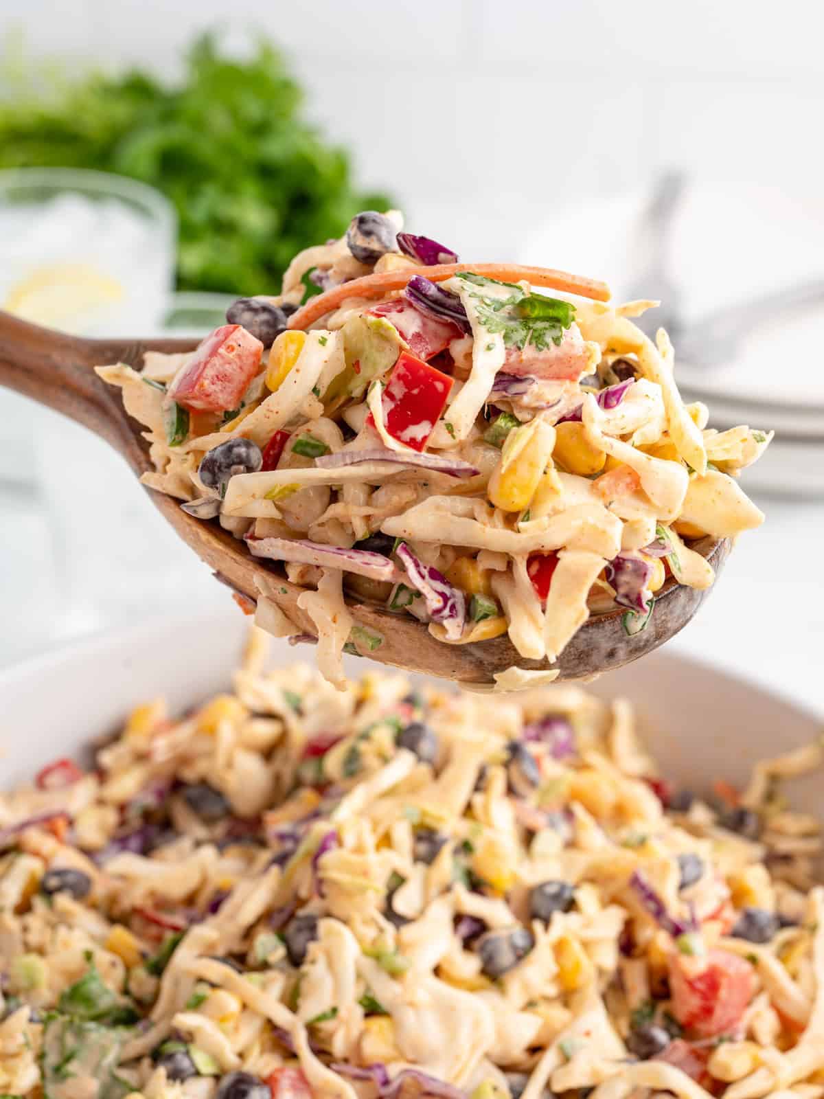 A wooden spoon lifting a serving of creamy taco coleslaw with black beans, corn, and red bell peppers over a full bowl.