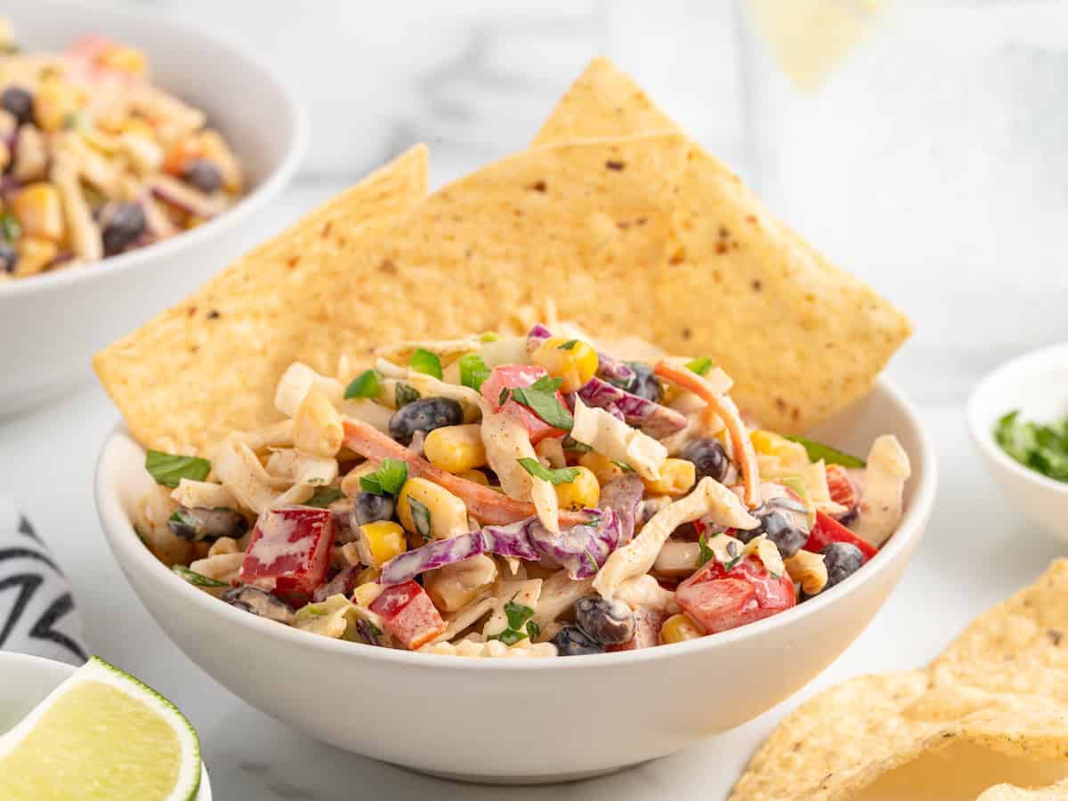 A bowl of taco coleslaw with chicken, black beans, corn, and peppers, served with tortilla chips and a lime wedge.