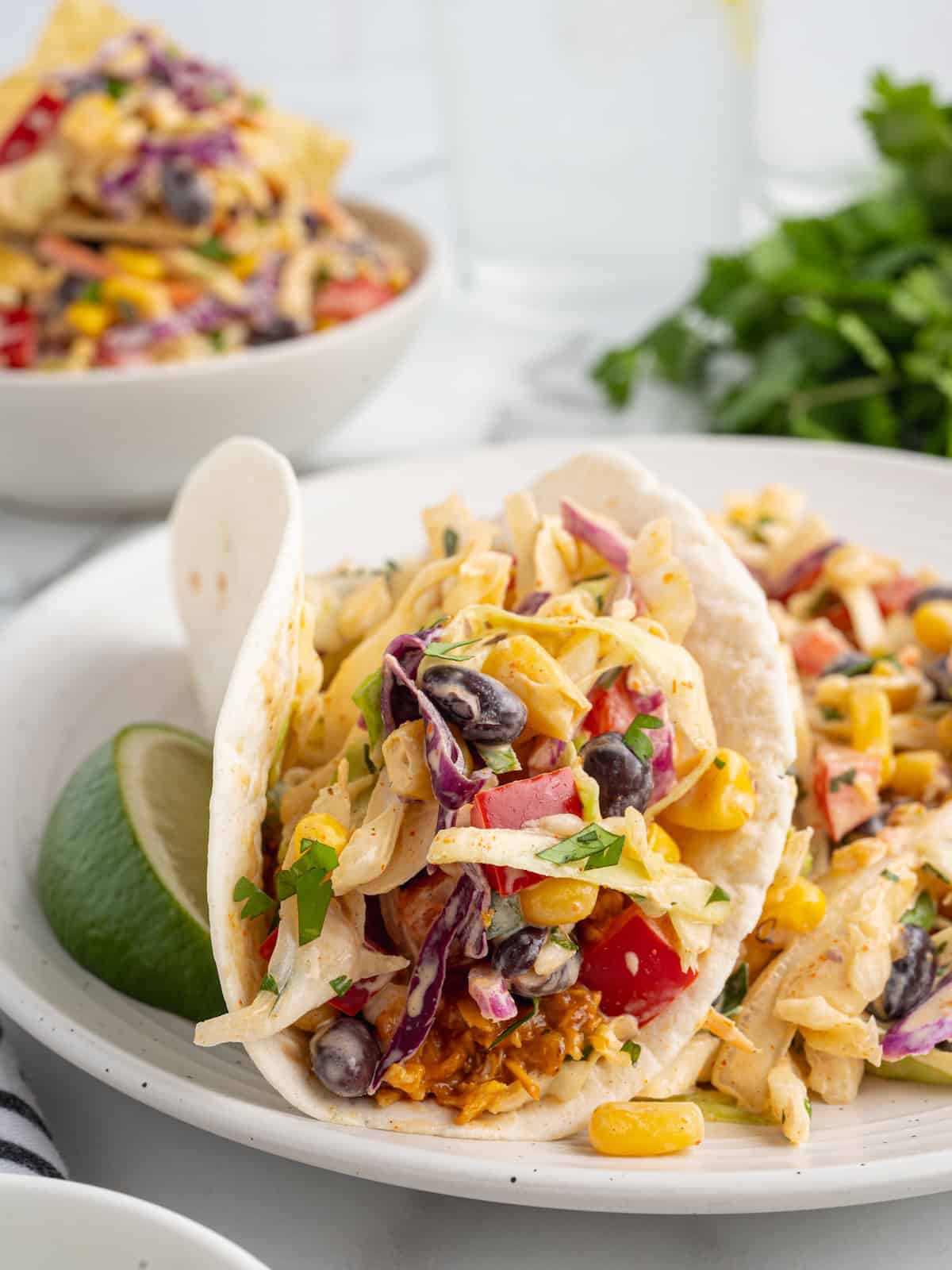 Two tacos in flour tortillas filled with black beans, corn, red bell pepper, and taco coleslaw, served with a lime wedge.