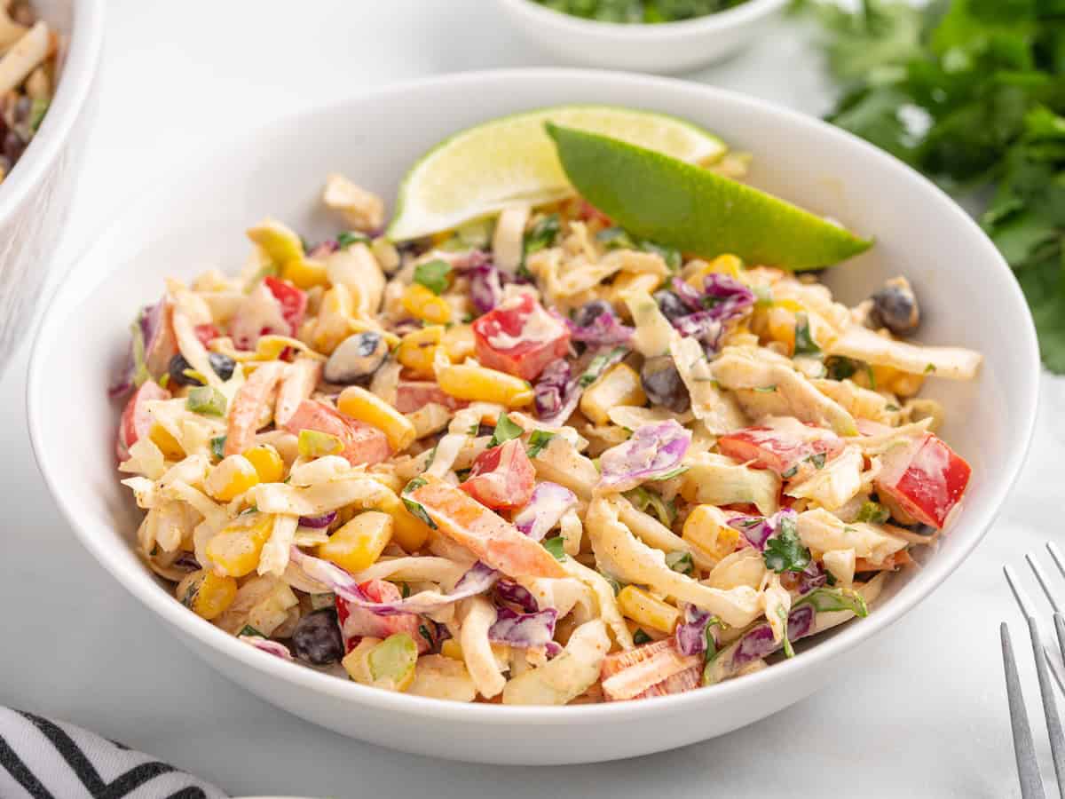 A bowl of creamy taco coleslaw mixed with corn, black beans, and diced red peppers, garnished with a lime wedge.