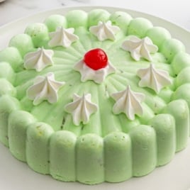 Whole seafoam salad molded in a circular shape, topped with dollops of whipped cream and a center maraschino cherry, surrounded by white plates and cherries.