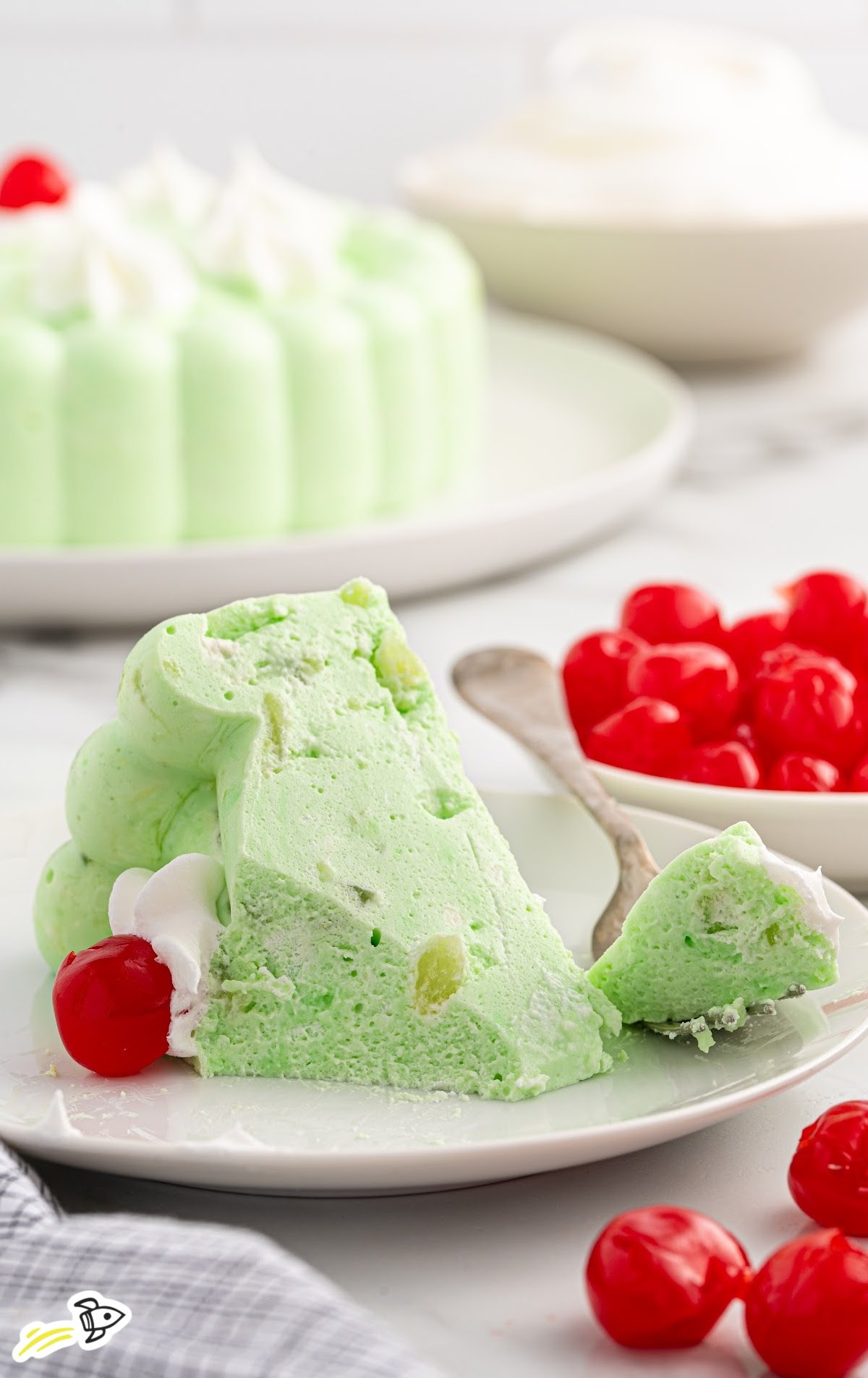 Slice of green seafoam salad on a plate with a spoon, showing creamy interior and chunks of pear, with cherries and the full mold in the background.