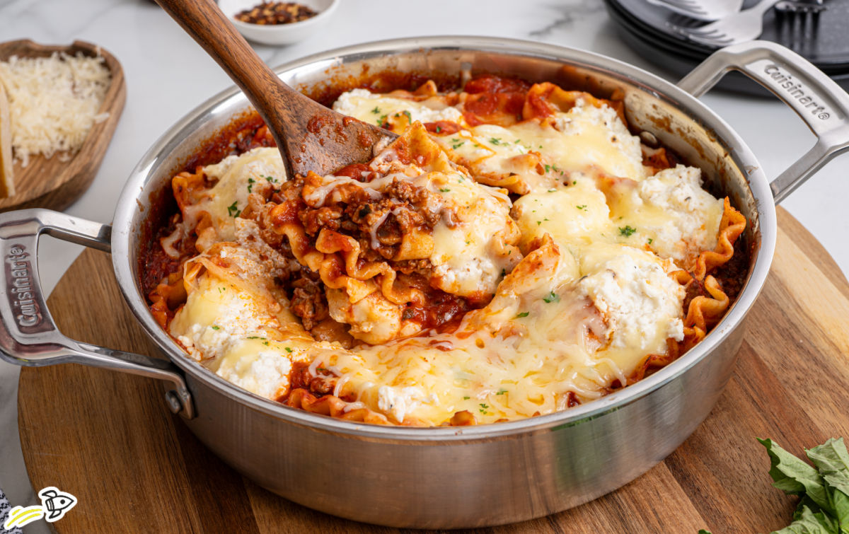 Skillet lasagna in a stainless steel pan with layers of wavy noodles, meat sauce, ricotta, and melted mozzarella, being scooped with a wooden spoon.