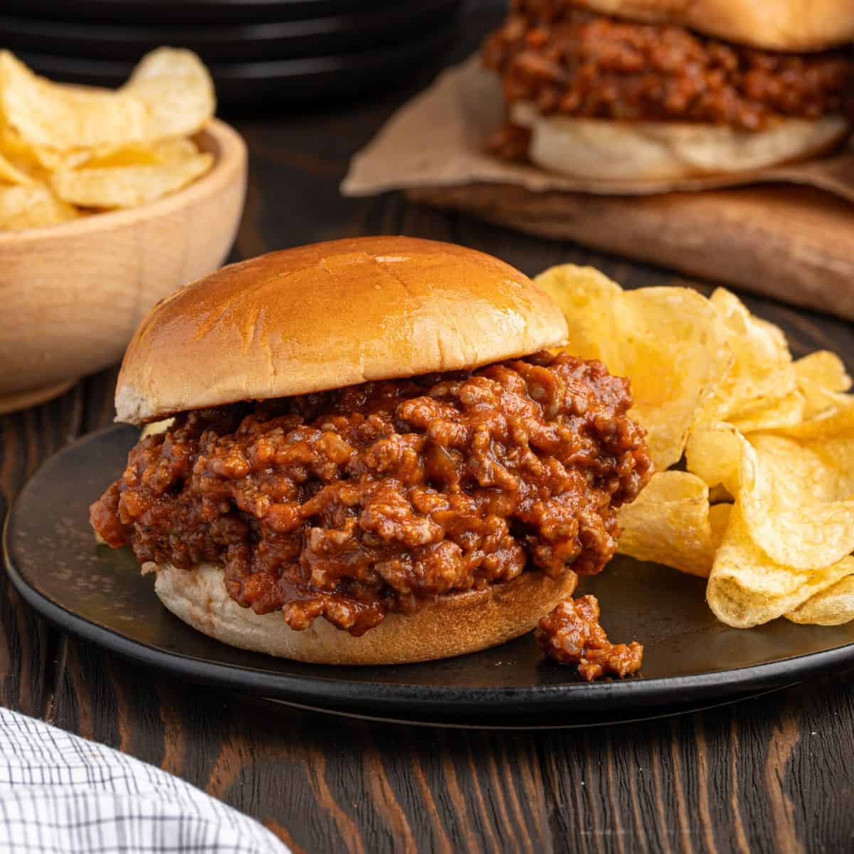 A classic Sloppy Joes sandwich filled with saucy ground meat sits on a black plate next to a pile of potato chips. A bowl of chips and another hearty Sloppy Joes sandwich are in the background.