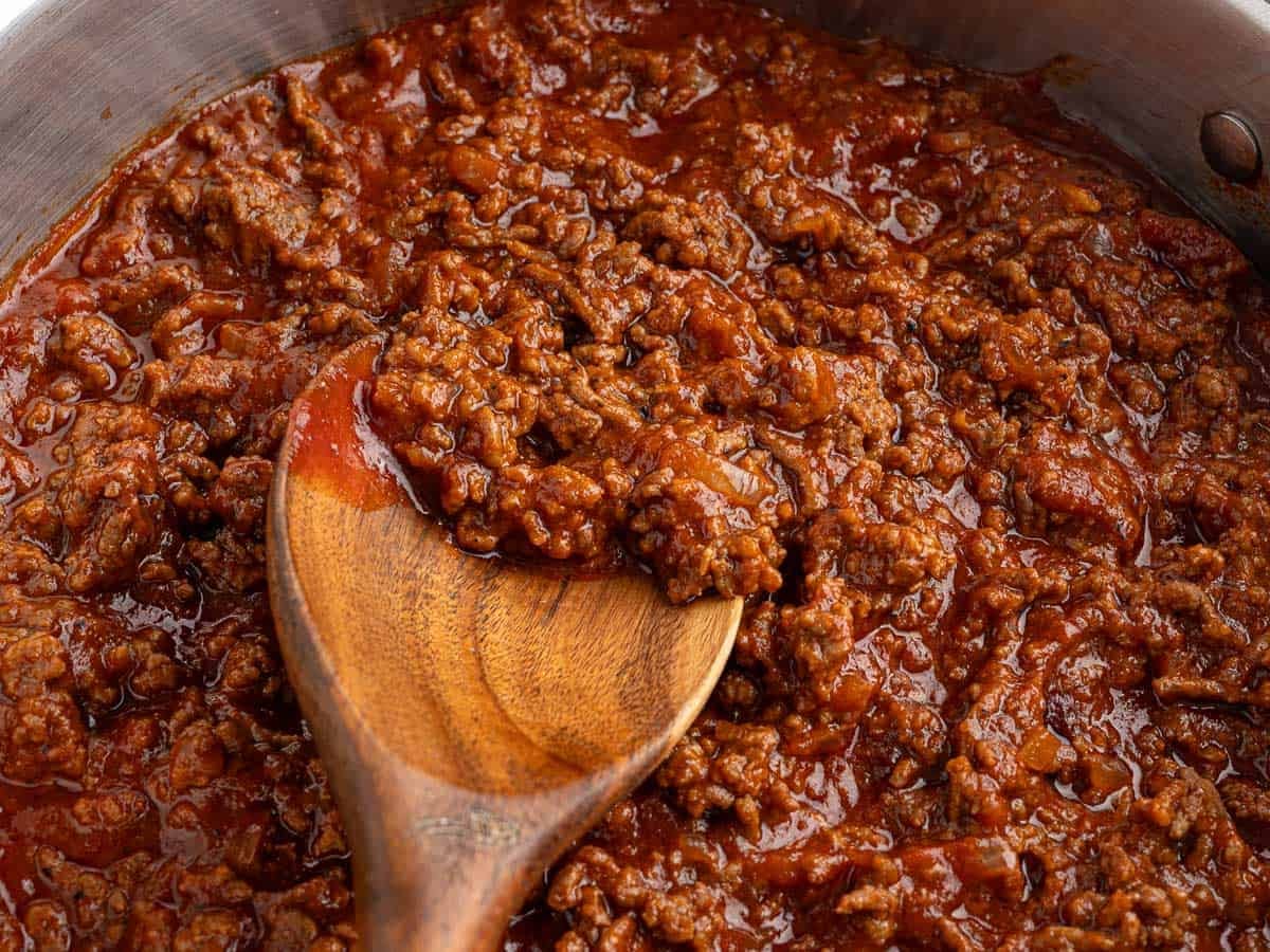 A close-up of a wooden spoon stirring thick, chunky ground beef in tomato sauce—perfect for making classic Sloppy Joes—inside a metal pan.