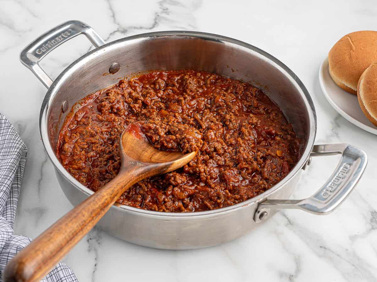 A stainless steel pan filled with savory Sloppy Joes mixture sits on a marble counter, with a wooden spoon resting inside. A plate with hamburger buns is visible to the right, ready to be piled high with the classic Sloppy Joes filling.