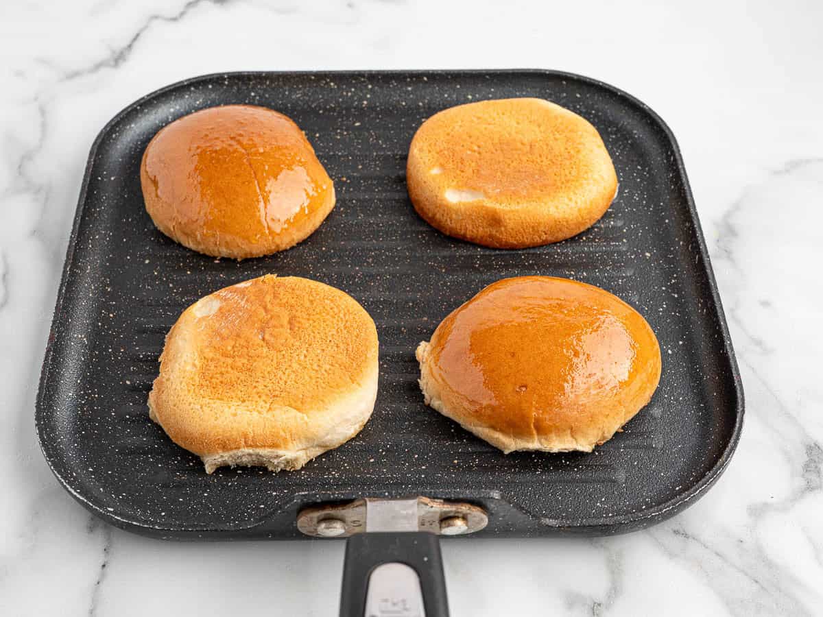 Four hamburger bun halves toasting on a square grill pan, perfect for Sloppy Joes, with two cut sides down and two rounded tops facing up, on a white marble surface.