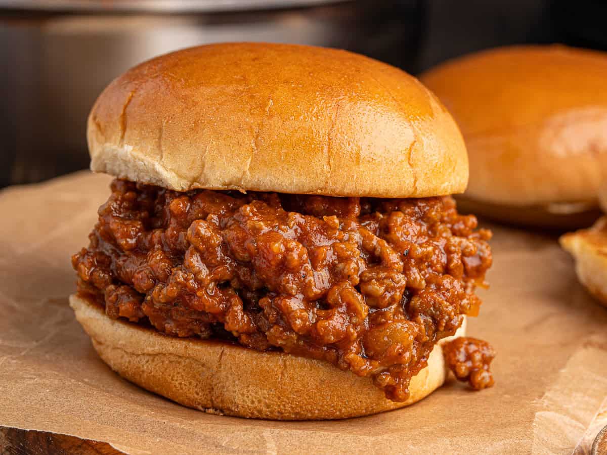 A classic Sloppy Joes sandwich filled with seasoned ground beef and rich tomato sauce sits on brown parchment paper, with another messy, irresistible sandwich in the background.