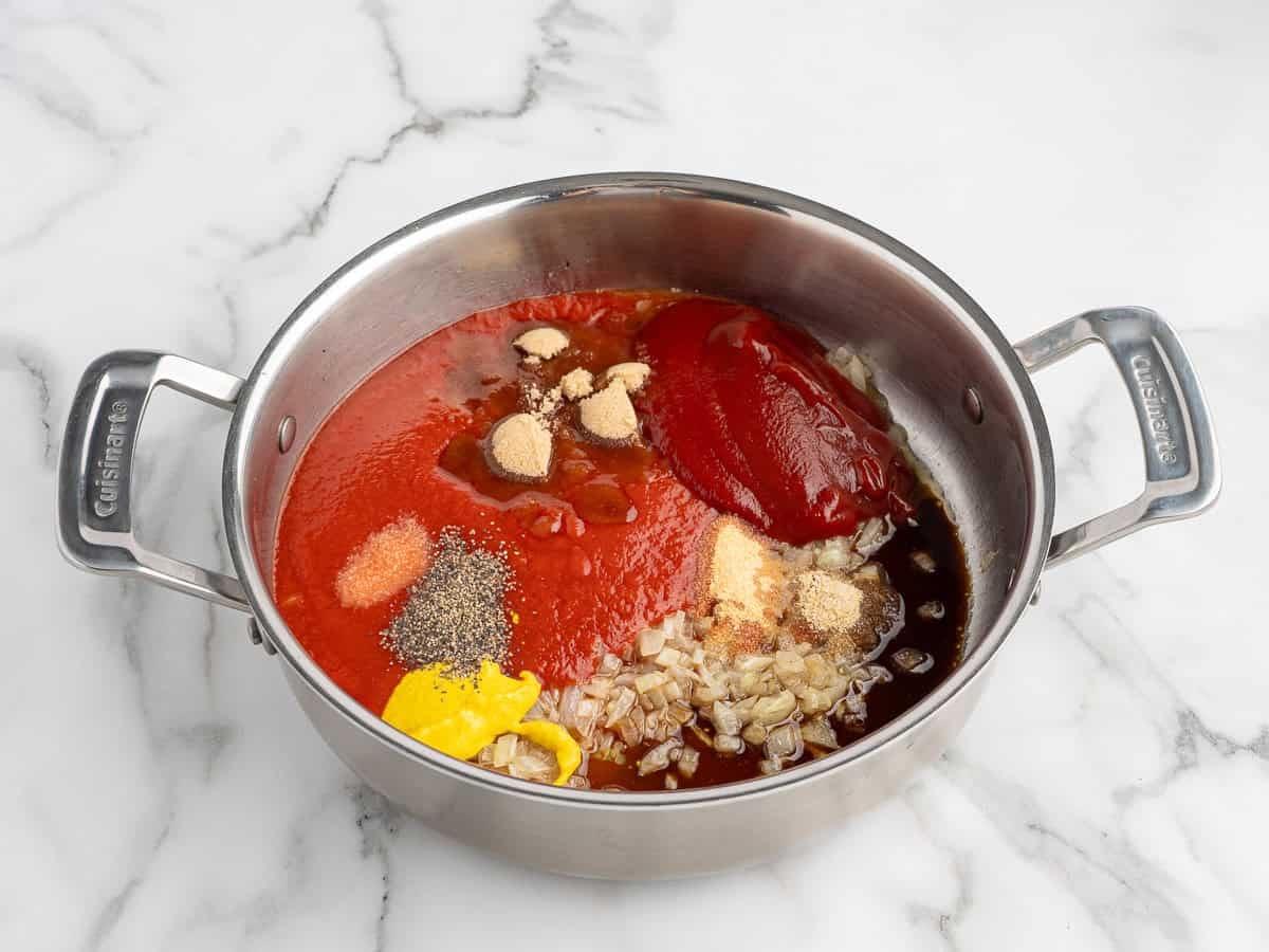 A stainless steel pot on a marble counter filled with chopped onions, ketchup, tomato paste, mustard, spices, and sauce ingredients for Sloppy Joes, ready to be mixed.