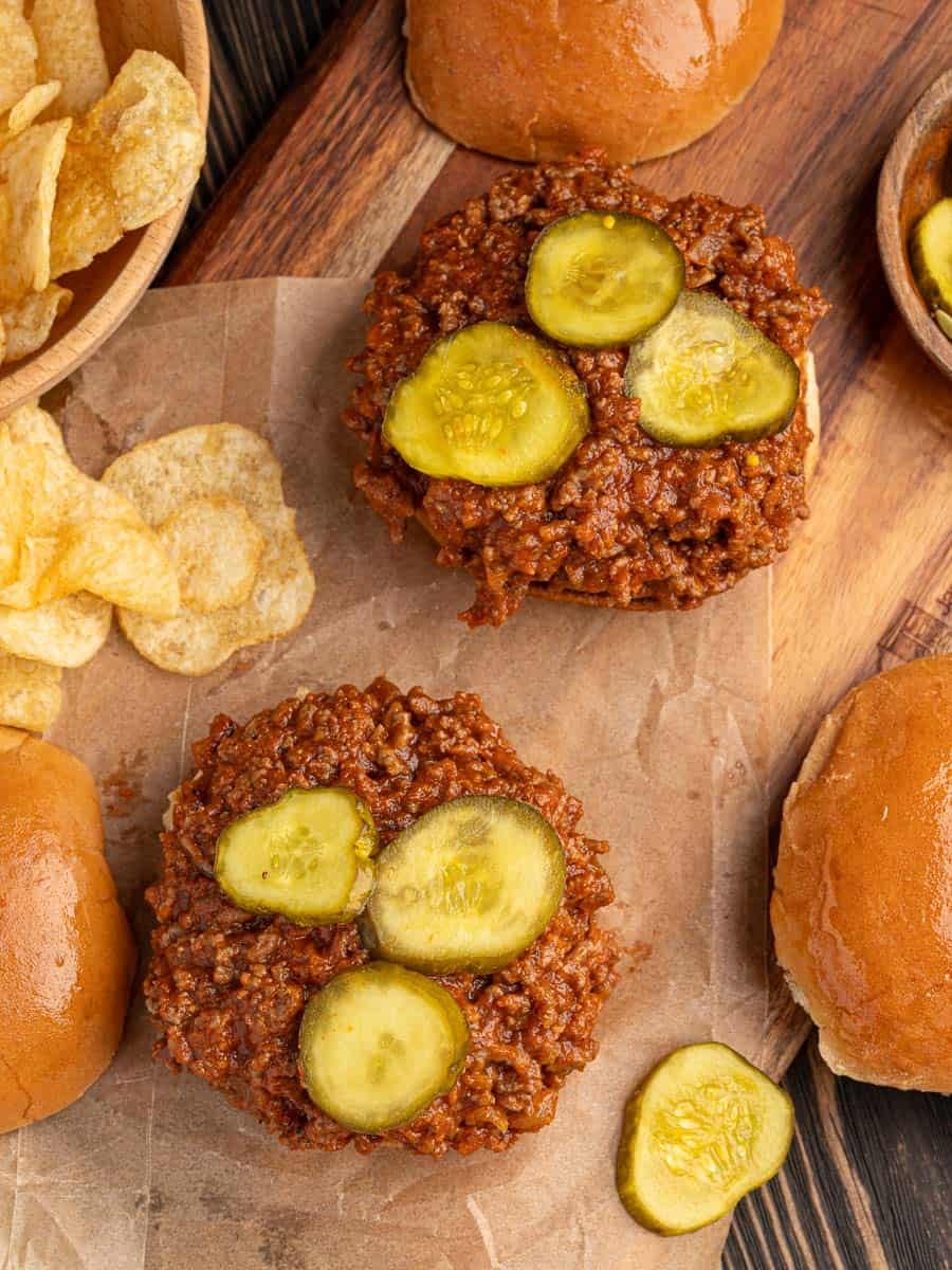 Two open-faced sloppy joe sandwiches, featuring classic Sloppy Joes filling and topped with sliced pickles, rest on parchment paper, surrounded by crispy potato chips and sandwich buns on a rustic wooden surface.