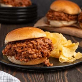 A plate featuring a classic Sloppy Joes sandwich with a side of crunchy potato chips sits on a wooden table, while more Sloppy Joes and a bowl of chips can be seen in the background.