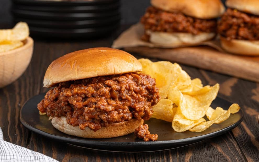 A plate featuring a classic Sloppy Joes sandwich with a side of crunchy potato chips sits on a wooden table, while more Sloppy Joes and a bowl of chips can be seen in the background.