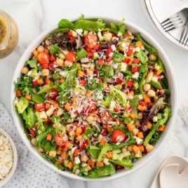 Top-down view of a large bowl of chopped salad with chickpeas, grape tomatoes, diced vegetables, mixed greens, and shredded cheese, surrounded by dressing, feta, and utensils.