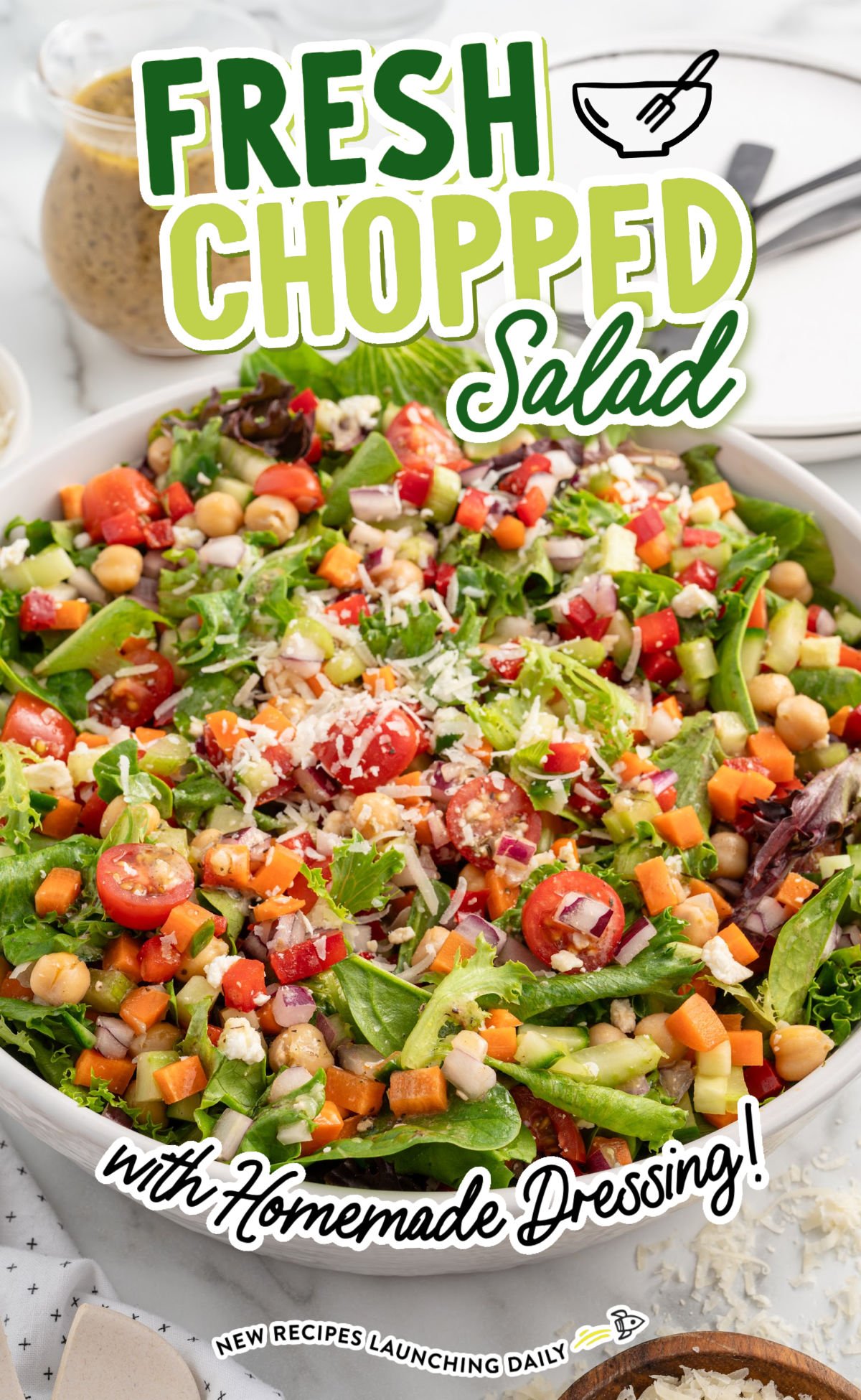 Vertical image of a fresh chopped salad in a white bowl with serving spoons, topped with shredded cheese and filled with chickpeas, tomatoes, greens, and diced veggies.