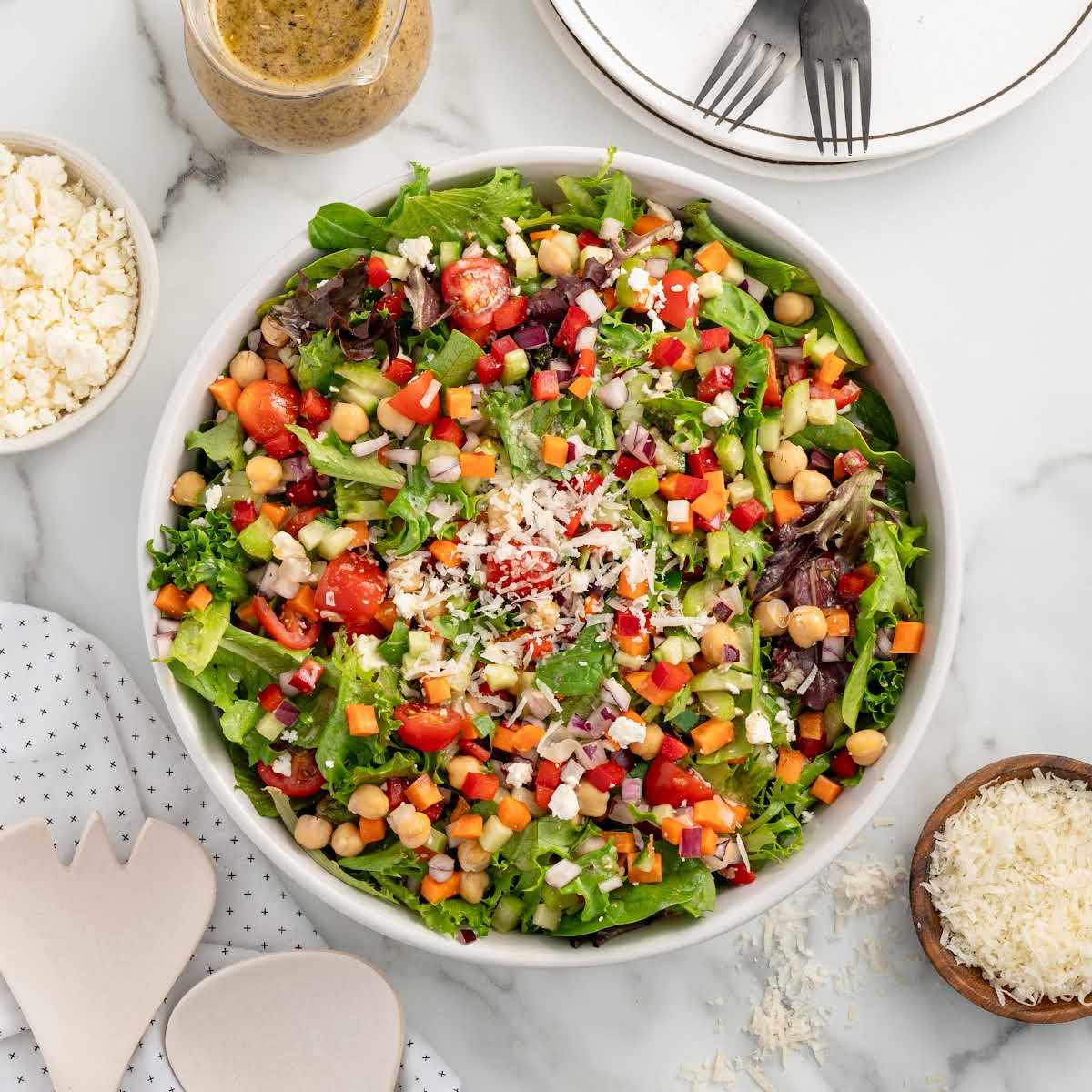 Top-down view of a fully assembled chopped salad with parmesan cheese and dressing on the side, set on a white surface with plates and forks nearby.