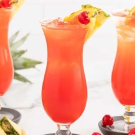 Three hurricane glasses filled with rum punch, garnished with pineapple, cherry, and orange slice.