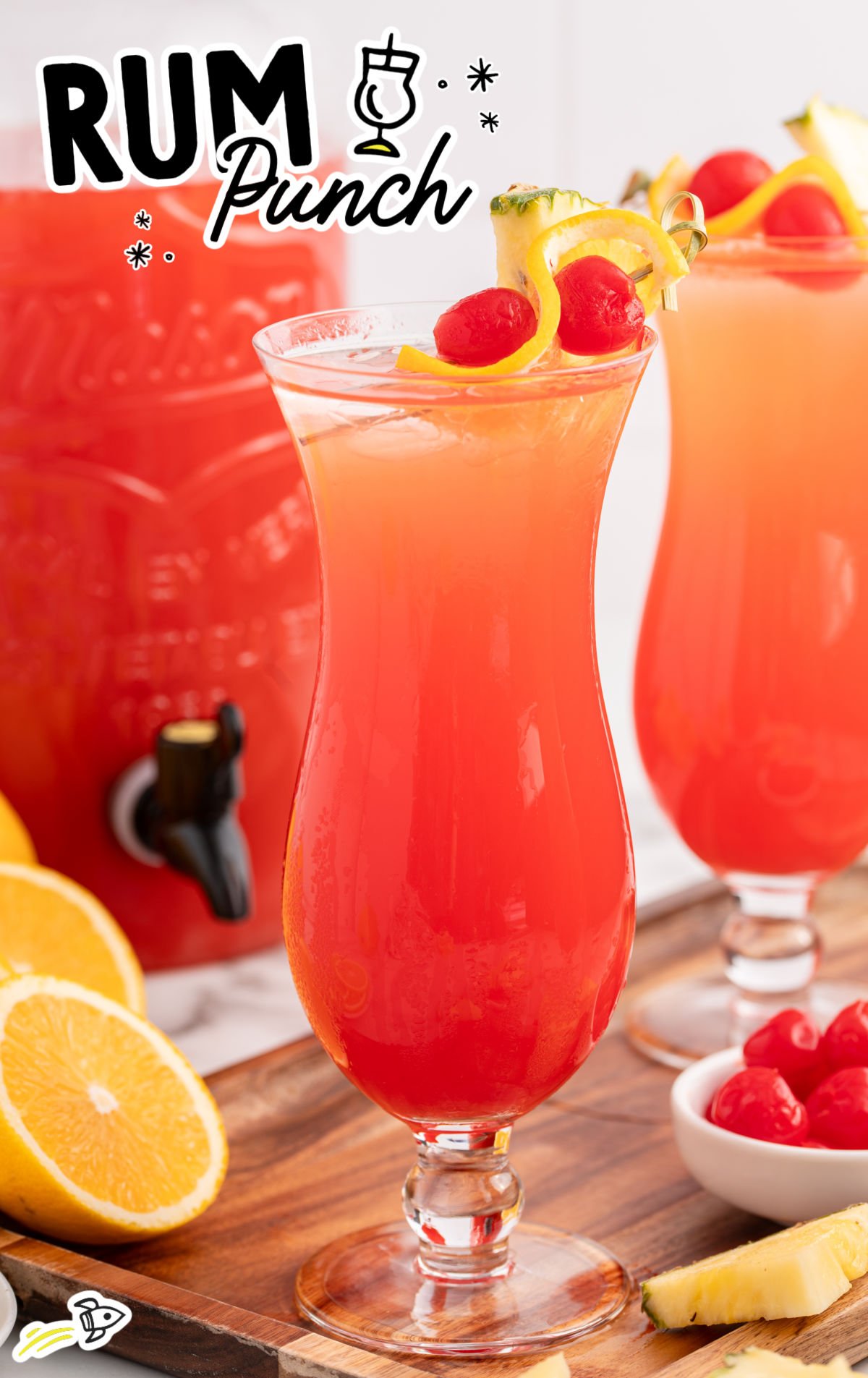 Close-up of a rum punch drink in a hurricane glass with a cherry and pineapple garnish, surrounded by tropical fruit.