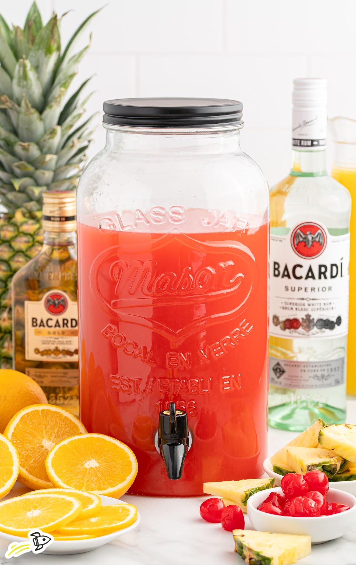 Large glass drink dispenser filled with rum punch, with rum bottles, pineapples, oranges, and cherries nearby.