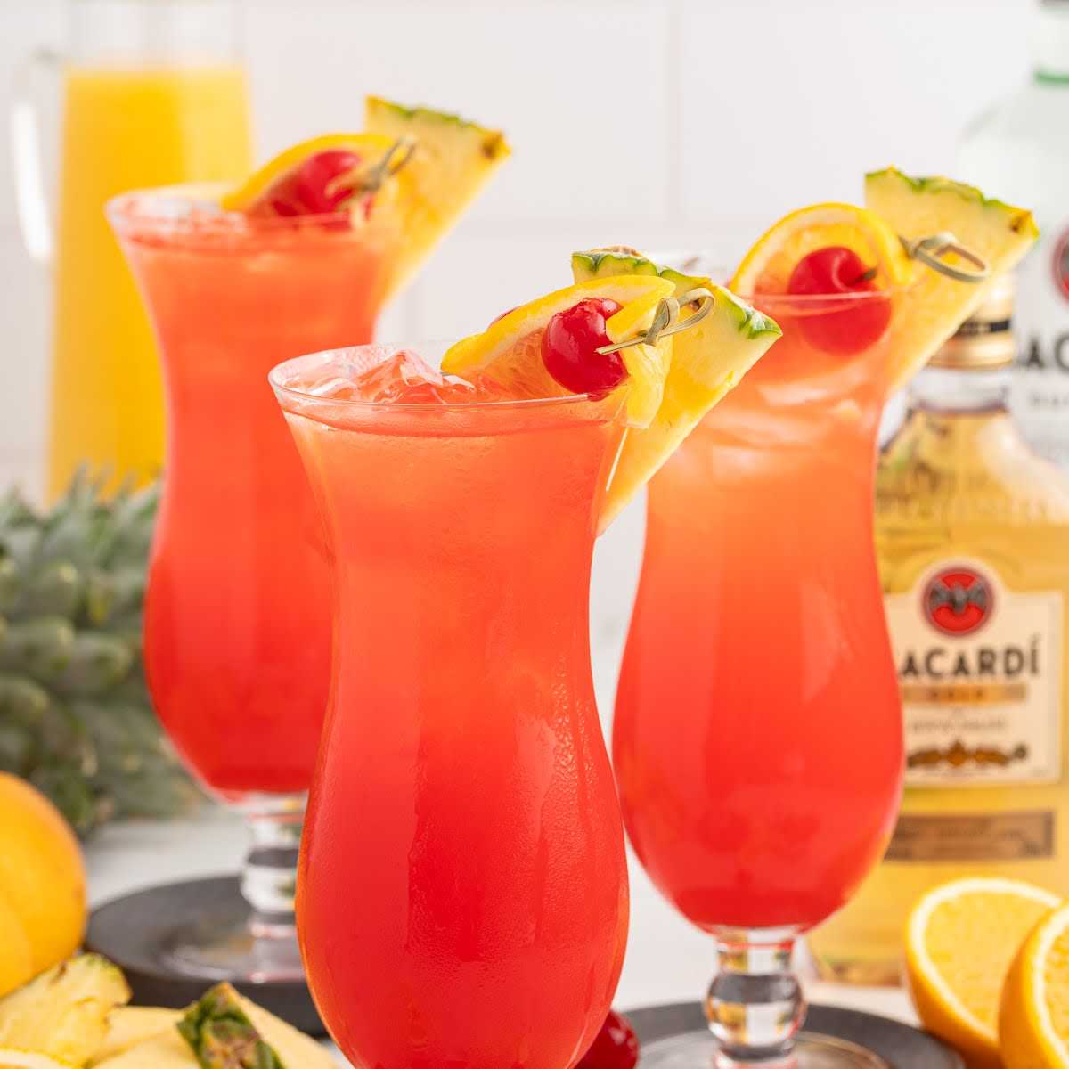 Three glasses of rum punch garnished with cherry, orange, and pineapple with bottles of Bacardi rum in the background.