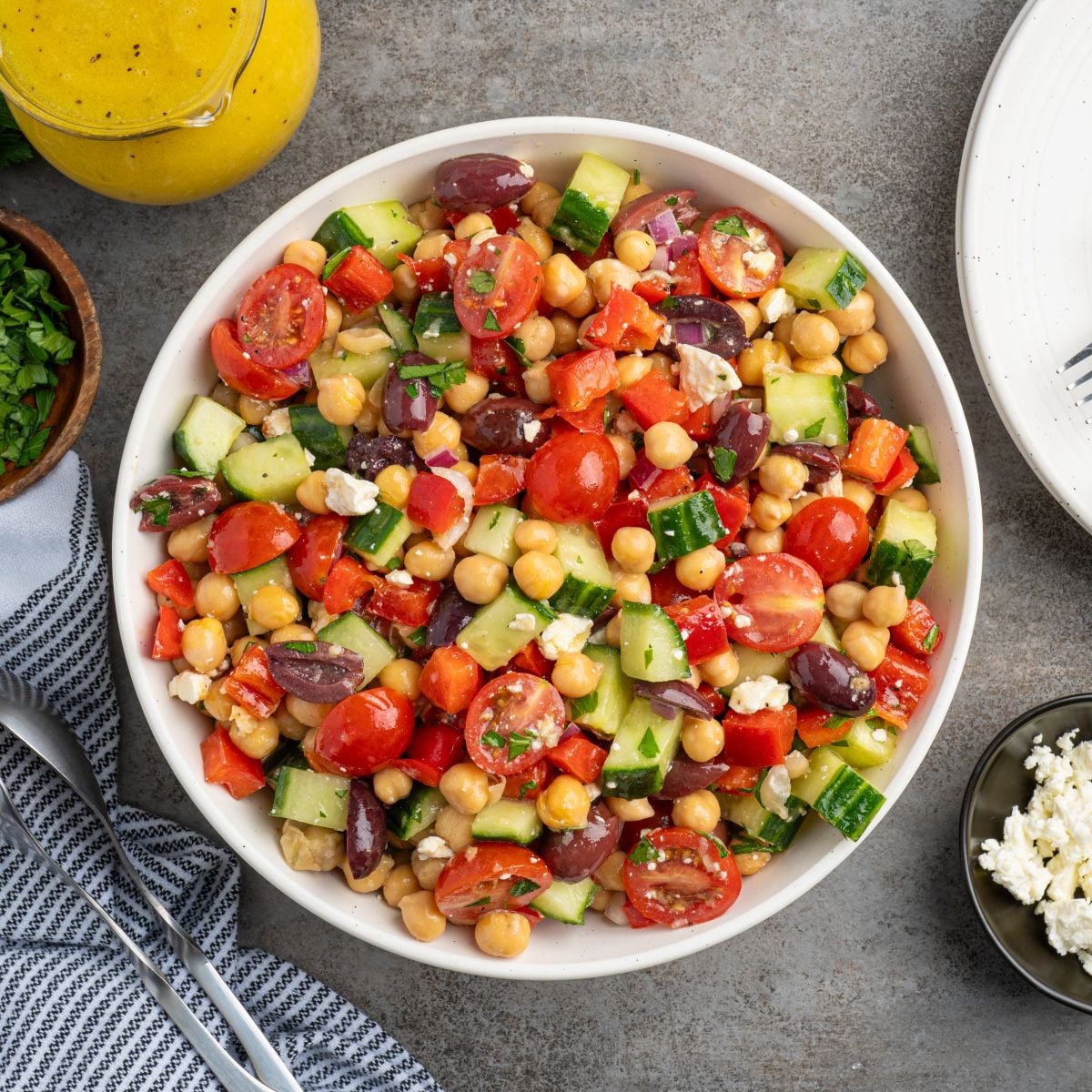 A bowl of Chickpea Salad with cherry tomatoes, cucumbers, red bell peppers, red onions, Kalamata olives, feta cheese, and parsley sits on a gray surface next to a striped napkin, cutlery, and small dishes.