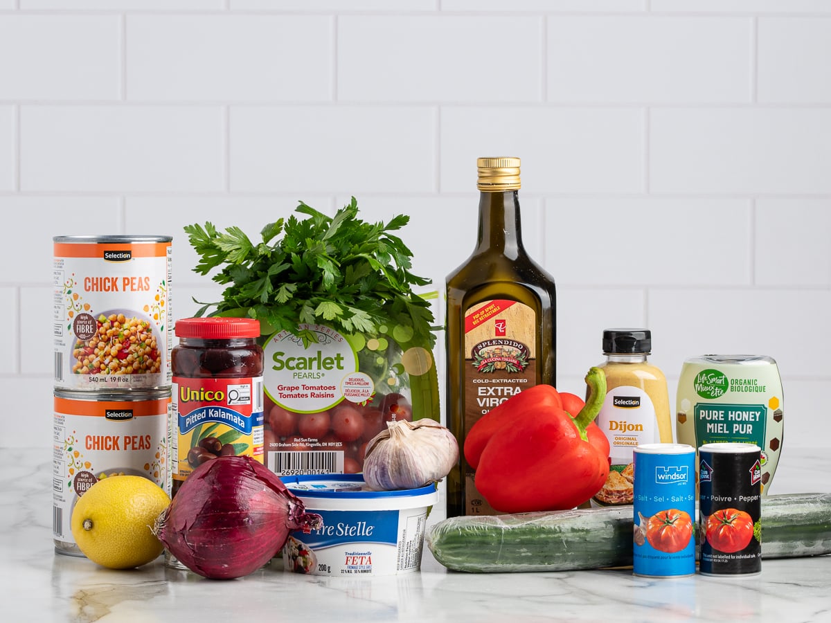 A variety of fresh and canned ingredients for Chickpea Salad on a white countertop, including chickpeas, parsley, feta, red onion, lemon, red pepper, cucumber, garlic, olive oil, Dijon mustard, honey, olives, salt, and pepper.