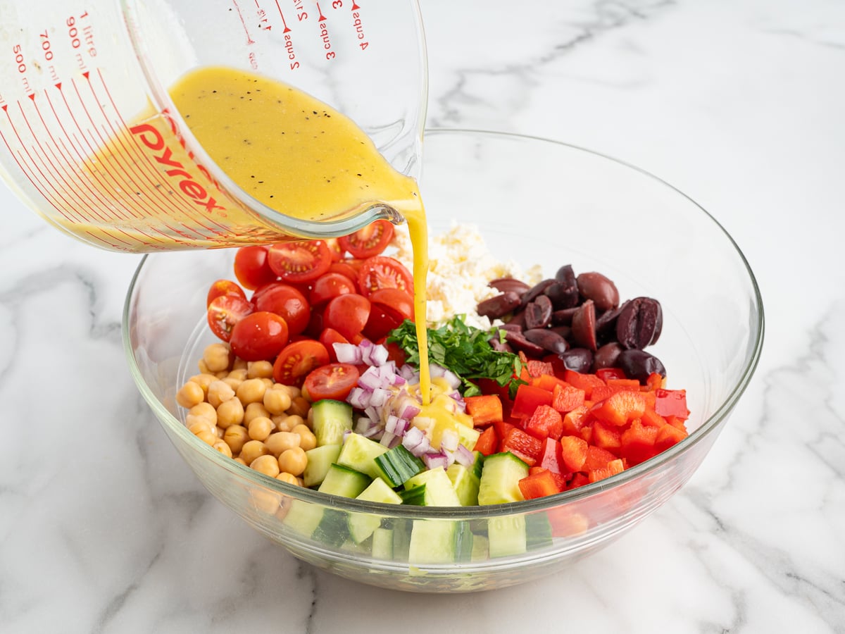 A measuring cup pours yellow dressing into a glass bowl filled with chopped cucumber, red onion, cherry tomatoes, bell pepper, olives, feta cheese, parsley, and hearty chickpeas for a vibrant Chickpea Salad on a marble countertop.