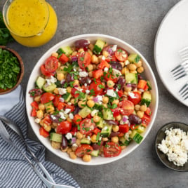 A bowl of vibrant Mediterranean Chickpea Salad with tomatoes, cucumbers, olives, and feta cheese on a gray surface, surrounded by fresh herbs, salad dressing, a small bowl of feta, plates, forks, and serving utensils.