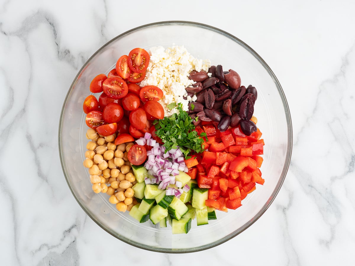 A glass bowl filled with separated ingredients for a Chickpea Salad: halved cherry tomatoes, sliced Kalamata olives, diced red bell pepper, chopped cucumber, chickpeas, diced red onion, crumbled feta cheese, and chopped parsley on a marble surface.