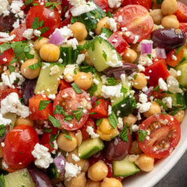 A close-up of a vibrant Chickpea Salad with cherry tomatoes, cucumber, red onion, Kalamata olives, red bell pepper, chopped parsley, and crumbled feta cheese in a white bowl.