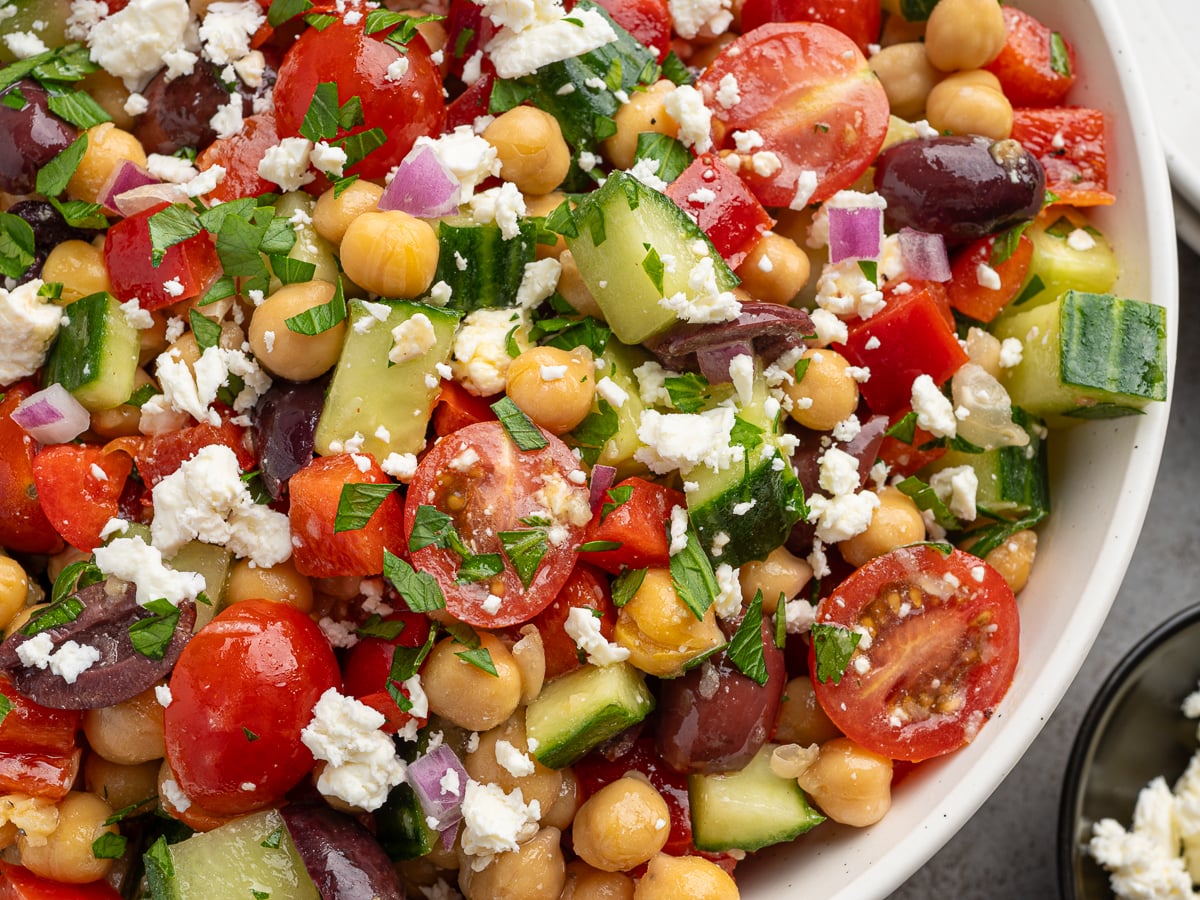 A close-up of a vibrant Chickpea Salad with cherry tomatoes, cucumber, red onion, Kalamata olives, red bell pepper, chopped parsley, and crumbled feta cheese in a white bowl.