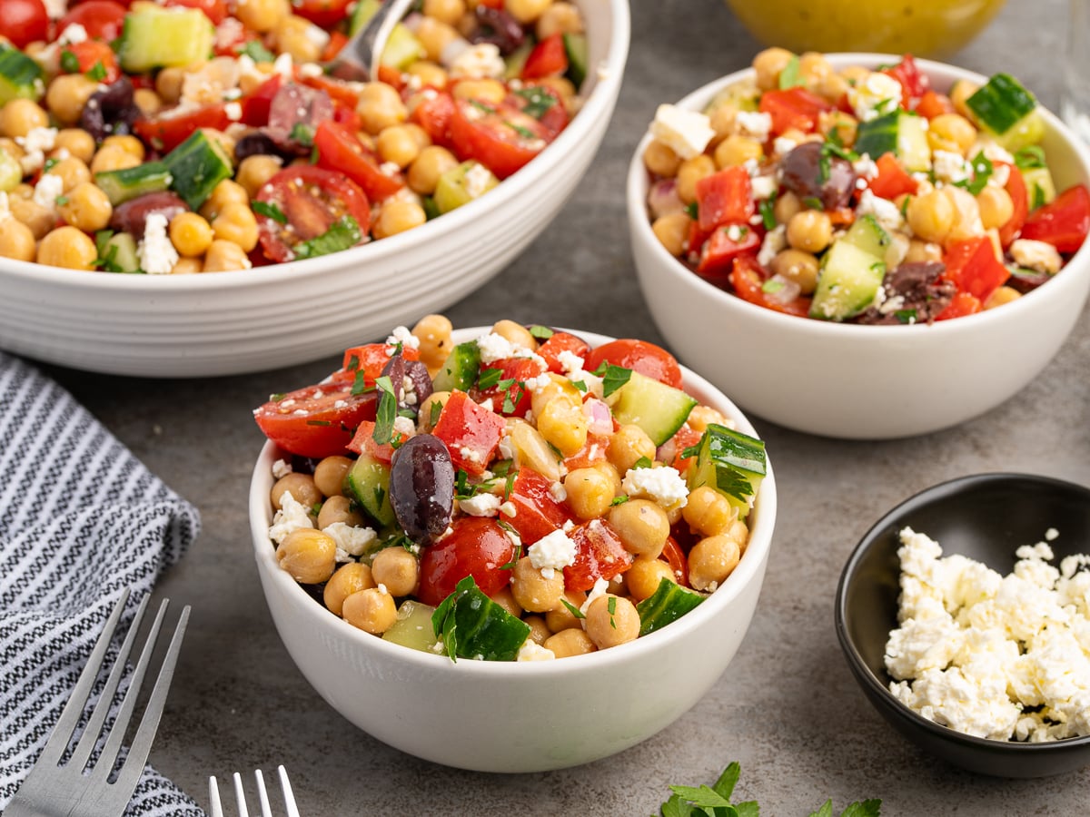 Bowls of Chickpea Salad with cherry tomatoes, cucumber, olives, parsley, and crumbled feta are arranged on a gray surface beside a small plate of feta and a striped napkin with forks.
