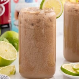 Three glasses of Dirty Dr Pepper with lime wedges and ice, set beside sliced limes, creamer, and Dr Pepper cans.