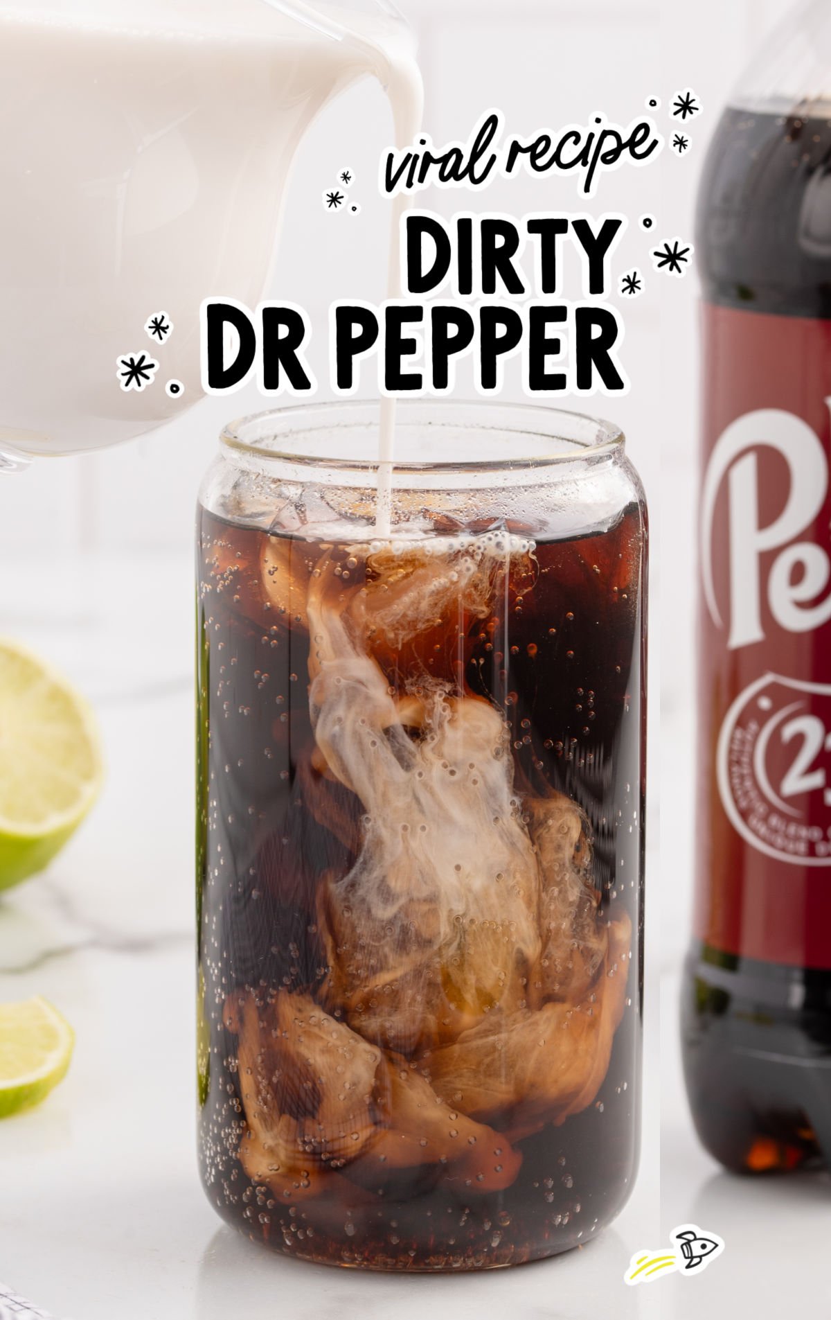 Creamer being poured into a glass of iced Dr Pepper with fresh limes and creamer bottle in the background.