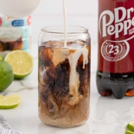 Creamer being poured into a glass of iced Dr Pepper with fresh limes and creamer bottle in the background.