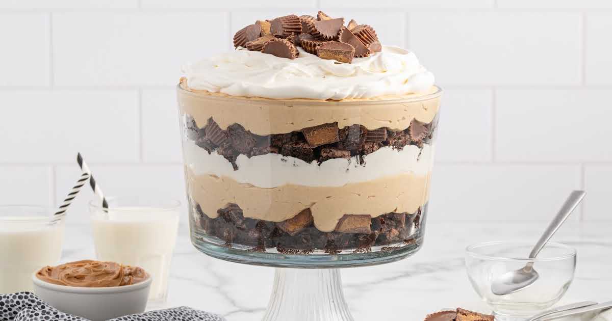 Layered peanut butter trifle in a glass bowl with whipped cream and chopped Reeseโs on top.