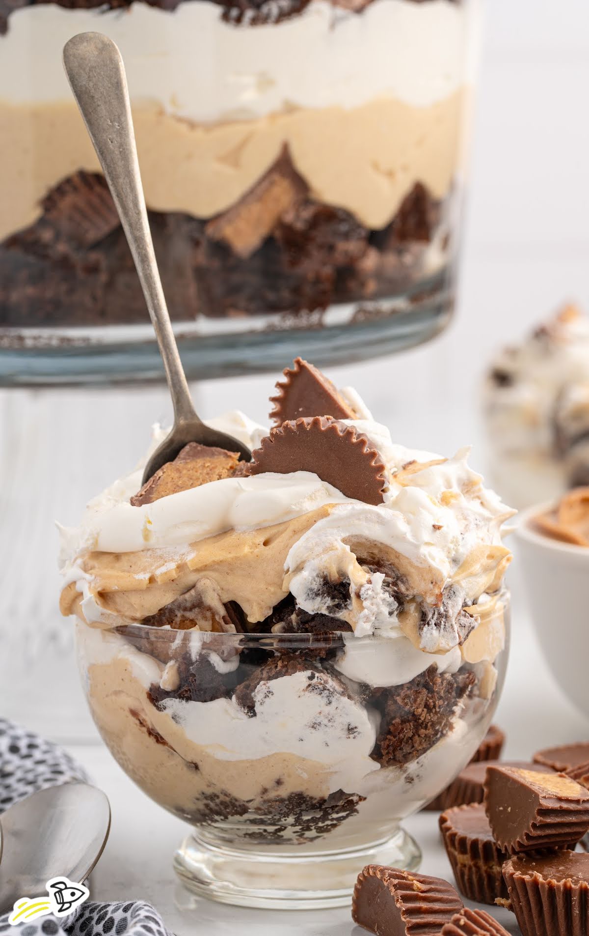 Close-up of a serving of peanut butter trifle in a small glass bowl with layers of brownie, whipped cream, peanut butter mousse, and mini Reese’s, with a spoon dipped in.