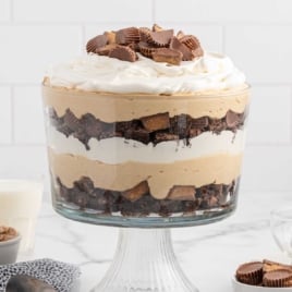 Front-facing view of layered peanut butter trifle topped with whipped cream and mini Reese’s, with milk and peanut butter in the background.