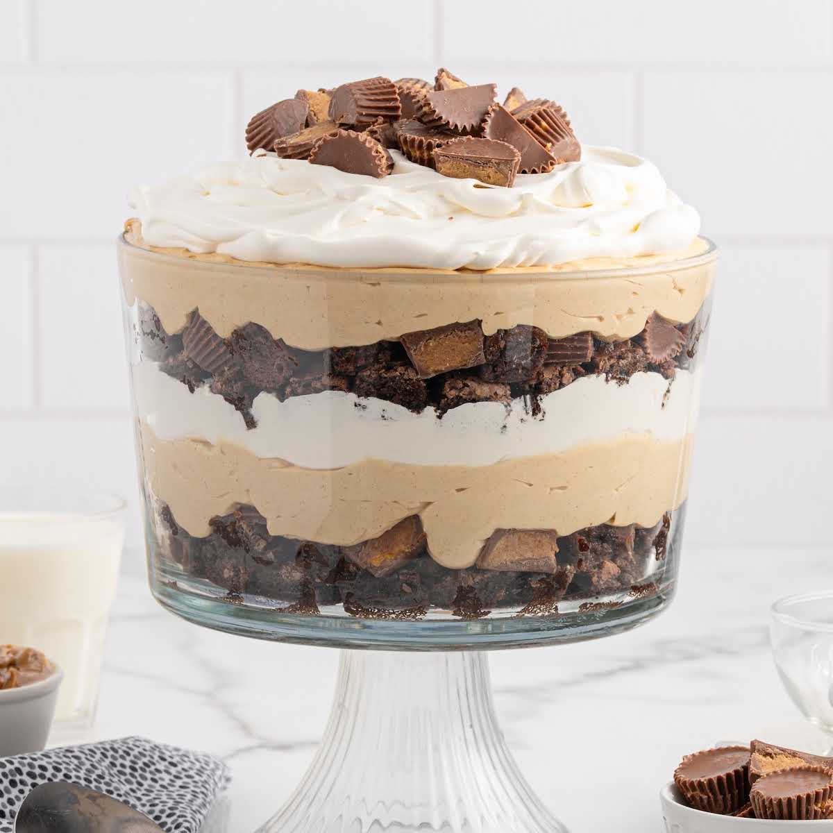 Front-facing view of layered peanut butter trifle topped with whipped cream and mini Reese’s, with milk and peanut butter in the background.
