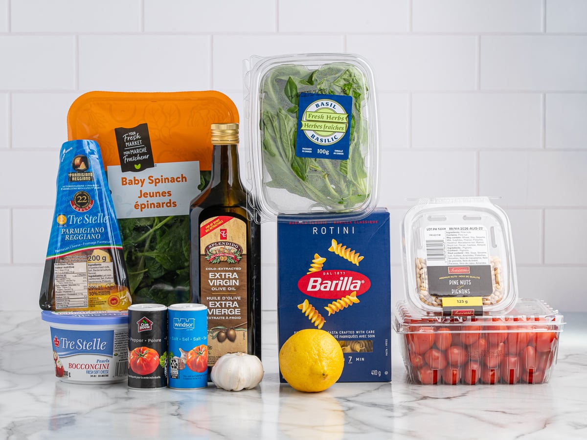 A variety of fresh and packaged ingredients for Pesto Pasta Salad on a marble counter, including baby spinach, basil, rotini pasta, grape tomatoes, bocconcini cheese, garlic, lemon, pine nuts, Parmesan cheese, olive oil, and canned tomatoes.