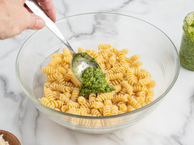A hand holds a spoon, adding green pesto to a glass bowl filled with cooked rotini pasta for a delicious Pesto Pasta Salad. A jar of pesto sits nearby on a marble countertop.