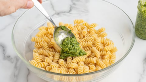 A hand holds a spoon, adding green pesto to a glass bowl filled with cooked rotini pasta for a delicious Pesto Pasta Salad. A jar of pesto sits nearby on a marble countertop.