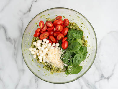 A glass bowl of Pesto Pasta Salad with spiral pasta, grape tomatoes, mozzarella balls, spinach leaves, and shredded parmesan cheese sits on a marble surface.