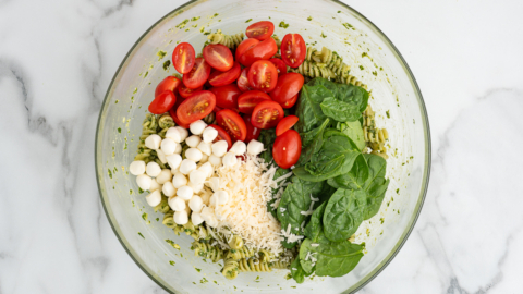 A glass bowl of Pesto Pasta Salad with spiral pasta, grape tomatoes, mozzarella balls, spinach leaves, and shredded parmesan cheese sits on a marble surface.