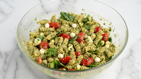 A glass bowl filled with Pesto Pasta Salad—rotini pasta mixed with green pesto, cherry tomatoes, fresh spinach, and small mozzarella balls—sits on a white marble surface.